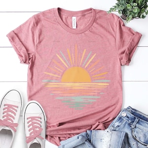 Op de afbeelding: Een heather mauve t-shirt met korte mouwen en een afbeelding van een gele zon die opkomt boven een horizon, met zonnestralen en waterreflecties in tinten geel, oranje en saliegroen. Het shirt heeft een ronde hals en opgerolde mouwen.