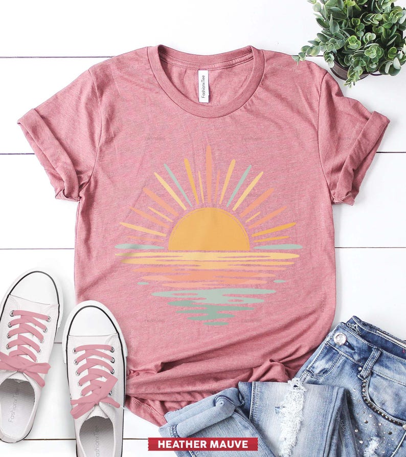 Op de afbeelding: Een heather mauve t-shirt met korte mouwen en een afbeelding van een gele zon die opkomt boven een horizon, met zonnestralen en waterreflecties in tinten geel, oranje en saliegroen. Het shirt heeft een ronde hals en opgerolde mouwen.