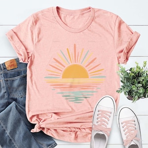 Op de afbeelding: Een perzikkleurig t-shirt met korte mouwen en een afbeelding van een opkomende zon boven water. De zon is geel met stralen in tinten oranje, geel en lichtblauw. De reflectie op het water weerspiegelt deze kleuren in horizontale banden.