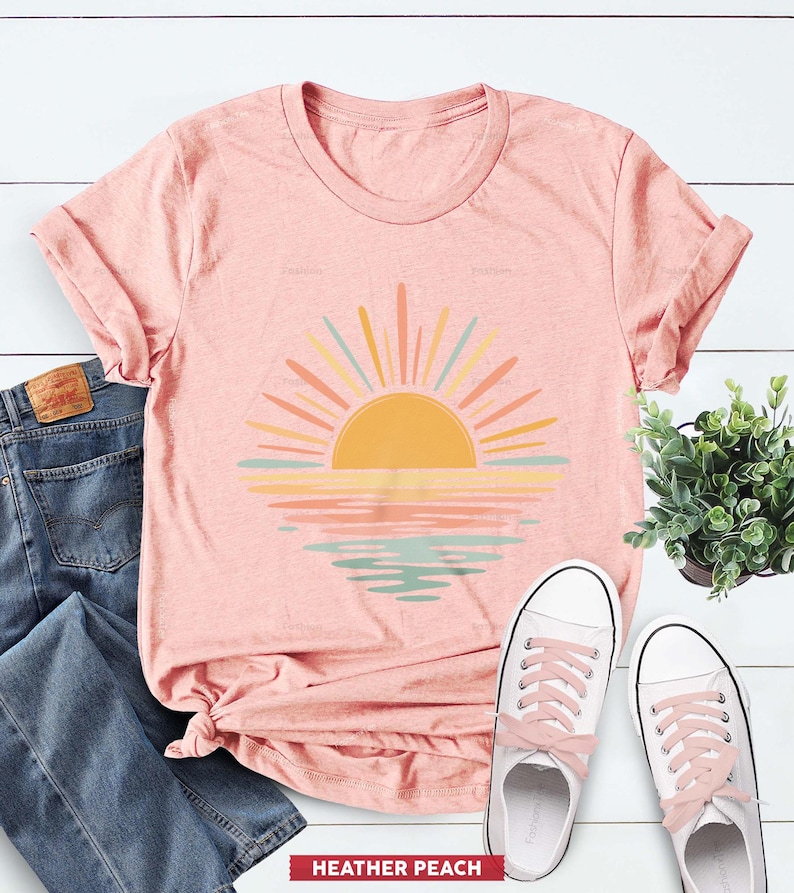 Op de afbeelding: Een perzikkleurig t-shirt met korte mouwen en een afbeelding van een opkomende zon boven water. De zon is geel met stralen in tinten oranje, geel en lichtblauw. De reflectie op het water weerspiegelt deze kleuren in horizontale banden.