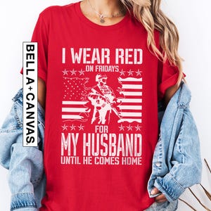 May include: A red short-sleeved t-shirt featuring a white graphic of a soldier standing in front of an American flag. The text reads I WEAR RED ON FRIDAYS FOR MY HUSBAND UNTIL HE COMES HOME. A vertical BELLA+CANVAS logo is printed on the left side.