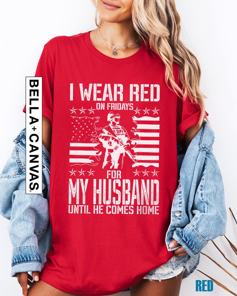 May include: A red short-sleeved t-shirt featuring a white graphic of a soldier standing in front of an American flag. The text reads I WEAR RED ON FRIDAYS FOR MY HUSBAND UNTIL HE COMES HOME. A vertical BELLA+CANVAS logo is printed on the left side.