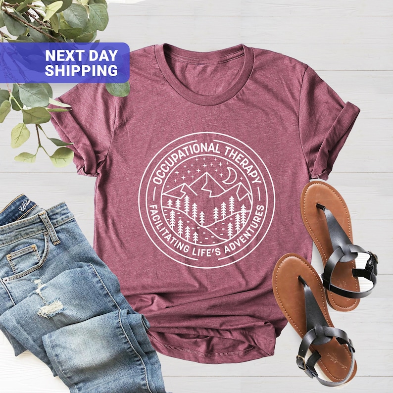 Op de afbeelding: Een maroon t-shirt met een wit grafisch ontwerp van een bergketen met een halve maan en sterren. De grafiek bevindt zich in een cirkel met de tekst "Occupational Therapy Facilitating Life's Adventures".