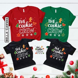 May include: A collection of red, green, black, and white shirts and a baby bodysuit featuring the text THE cookie CREW. The graphic includes illustrations of a gingerbread person, a Christmas tree, a heart, and ornaments with white dots.