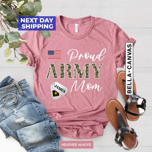 May include: A heather mauve t-shirt with the text "Proud Army Mom" in white. The word "Army" is in camouflage print. A small American flag is in the top left corner. A silver dog tag with the name "James" is hanging from a chain.