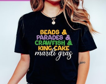 May include: A black short-sleeved t-shirt featuring the text BEADS & PARADES & CRAWFISH & KING CAKE mardi gras in yellow, purple, and green lettering. The shirt has a crew neckline and is worn with dark blue denim jeans.