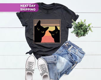 May include: A black short-sleeve t-shirt featuring a graphic of two black cats against a sunset-colored background with horizontal lines. The shirt is paired with blue denim jeans and a pair of white canvas sneakers with gray laces.