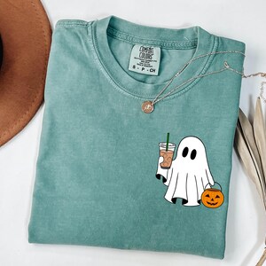 May include: A seafoam green t-shirt with a graphic of a ghost holding a coffee drink and a jack-o'-lantern. The shirt has a crew neck and short sleeves. A silver necklace with a pendant is draped across the shirt.