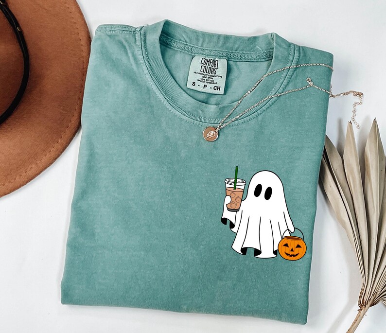 May include: A seafoam green t-shirt with a graphic of a ghost holding a coffee drink and a jack-o'-lantern. The shirt has a crew neck and short sleeves. A silver necklace with a pendant is draped across the shirt.