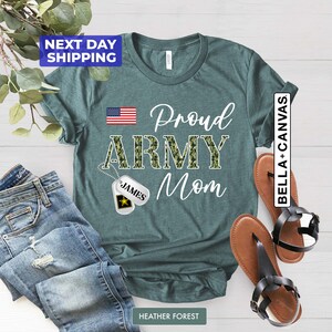 May include: Teal t-shirt with the text "Proud Army Mom" in white. The word "Army" is in camouflage print. A small American flag is in the top left corner. A silver dog tag with the name "James" is hanging from a chain.
