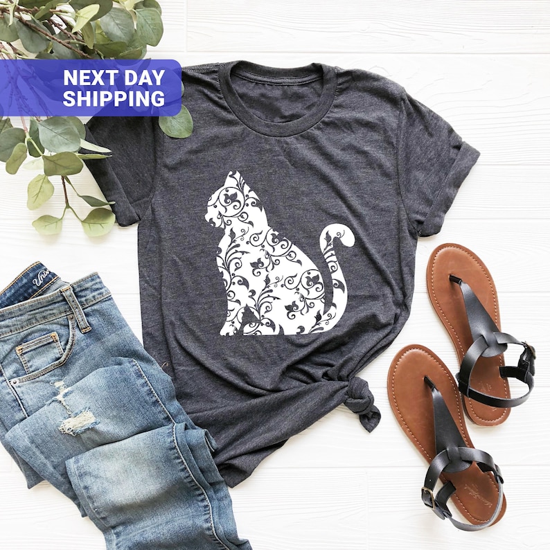 Puede incluir: Una camiseta gris oscuro con una silueta blanca de un gato con un patr&oacute;n floral. El gato est&aacute; sentado con la cola enroscada alrededor de su cuerpo. La camiseta est&aacute; colocada sobre una superficie blanca con un par de sandalias marrones y unos vaqueros azules.