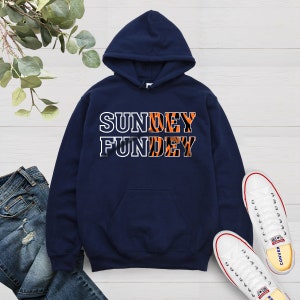 May include: A navy blue hoodie with a white pocket and the text "SUNDEY FUNDEY" in white letters with an orange and black tiger stripe pattern.
