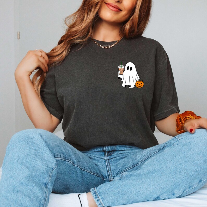May include: Dark gray t-shirt featuring a cartoon ghost holding a coffee drink and a jack-o'-lantern. The ghost is white with black eyes and a mouth. The jack-o'-lantern is orange with a carved face. The shirt is a casual style.