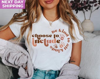 Może przedstawiać: Biały t-shirt z krótkim rękawem z brązowym i brzoskwiniowym napisem "choose to include" oraz "be kind to all kinds" ułożonym w kształt pętli nieskończoności, ozdobiony małymi grafikami odcisków dłoni z przodu ubrania.