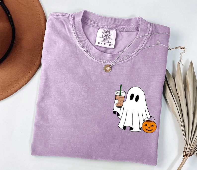 May include: A lavender Comfort Colors t-shirt featuring a white ghost graphic holding a coffee drink and a jack-o'-lantern. The shirt has a crew neck and short sleeves. A silver necklace with a pendant is also visible.
