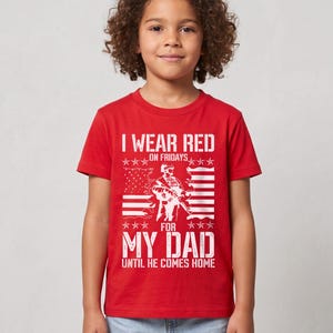 May include: A red short-sleeved t-shirt featuring a white graphic print of a soldier silhouette, a stylized American flag, and the text: I WEAR RED ON FRIDAYS FOR MY DAD UNTIL HE COMES HOME. The design includes star accents above and below the soldier.