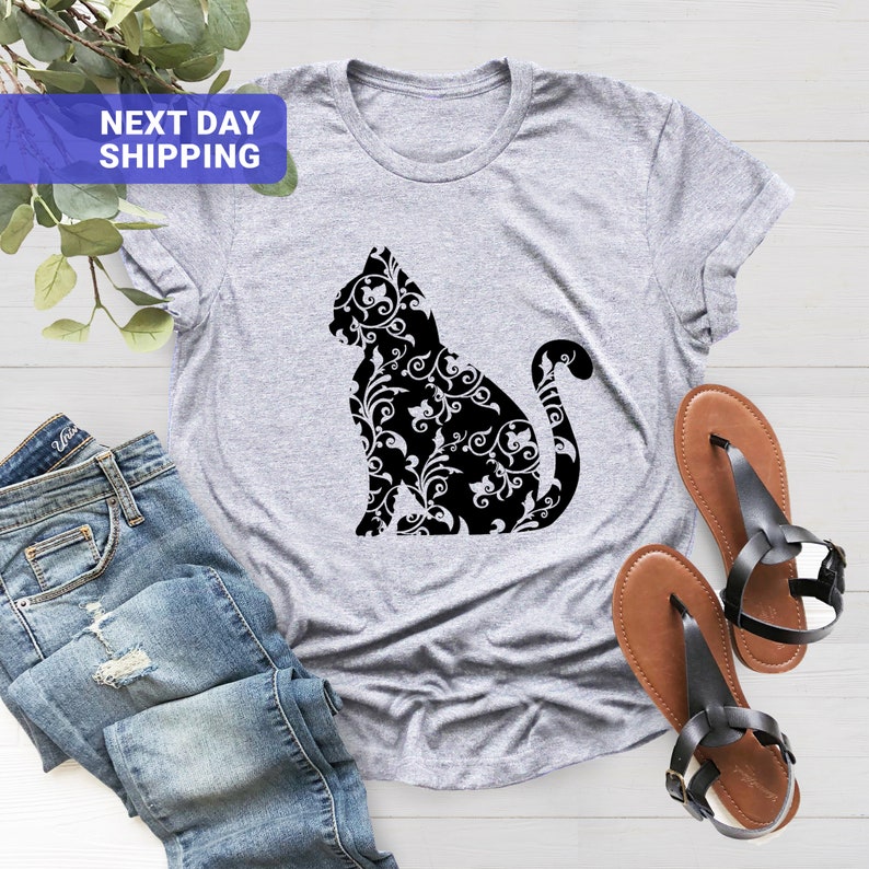 Puede incluir: Una camiseta gris con una silueta negra de un gato con un patr&oacute;n floral. El gato est&aacute; sentado con la cola enroscada alrededor de su cuerpo.
