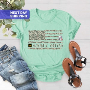 May include: A light green t-shirt featuring a camouflage American flag design and the text "Army Mom". A small tag with the word "David" is hanging from the flag.