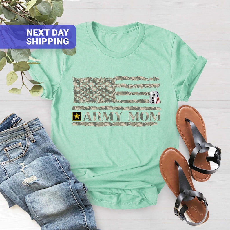 May include: A light green t-shirt featuring a camouflage American flag design and the text "Army Mom". A small tag with the word "David" is hanging from the flag.