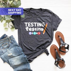 May include: A dark gray short-sleeve t-shirt featuring the white text "TESTING testing" and a graphic of a pencil next to the equation "1 + 2 = 3" with the numbers inside colored circles of purple, light blue, and red.