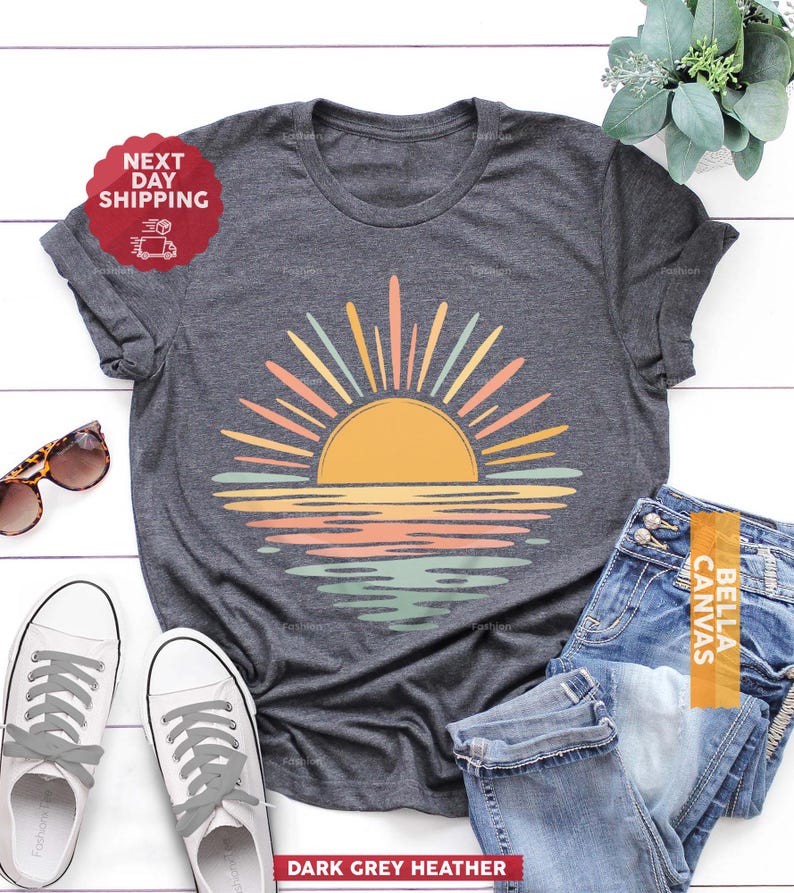 Op de afbeelding: Een donkergrijs gem&ecirc;leerd t-shirt met een afbeelding van een gestileerde zon die boven het water uitkomt. De zon is geel met stralende stralen in tinten perzik, geel en lichtgroen. De waterreflectie onder de zon gebruikt hetzelfde kleurenpalet.