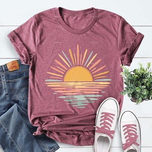 Op de afbeelding: Een bordeauxrood gem&ecirc;leerd T-shirt met korte mouwen en een afbeelding van een gele zon die opkomt boven een horizon, met zonnestralen en waterreflecties in tinten geel, perzik en lichtblauw. Het shirt wordt getoond met een blauwe spijkerbroek en sneakers.
