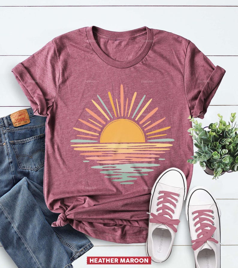 Op de afbeelding: Een bordeauxrood gem&ecirc;leerd T-shirt met korte mouwen en een afbeelding van een gele zon die opkomt boven een horizon, met zonnestralen en waterreflecties in tinten geel, perzik en lichtblauw. Het shirt wordt getoond met een blauwe spijkerbroek en sneakers.