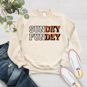 May include: A beige sweatshirt with the text "SUNDAY FUNDEY" printed on it. The letters are white with an orange and black tiger stripe pattern.