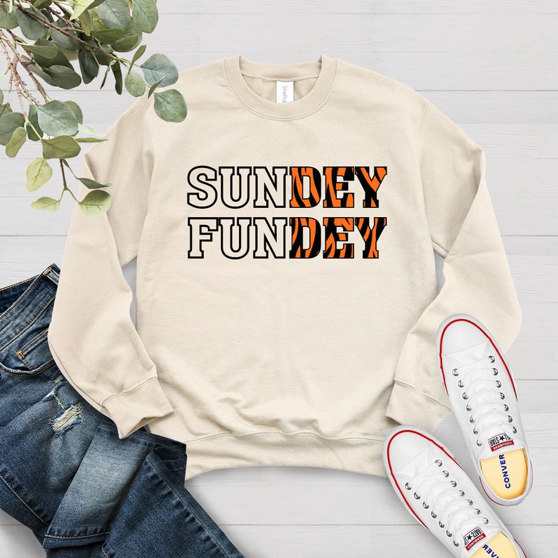 May include: A beige sweatshirt with the text "SUNDAY FUNDEY" printed on it. The letters are white with an orange and black tiger stripe pattern.