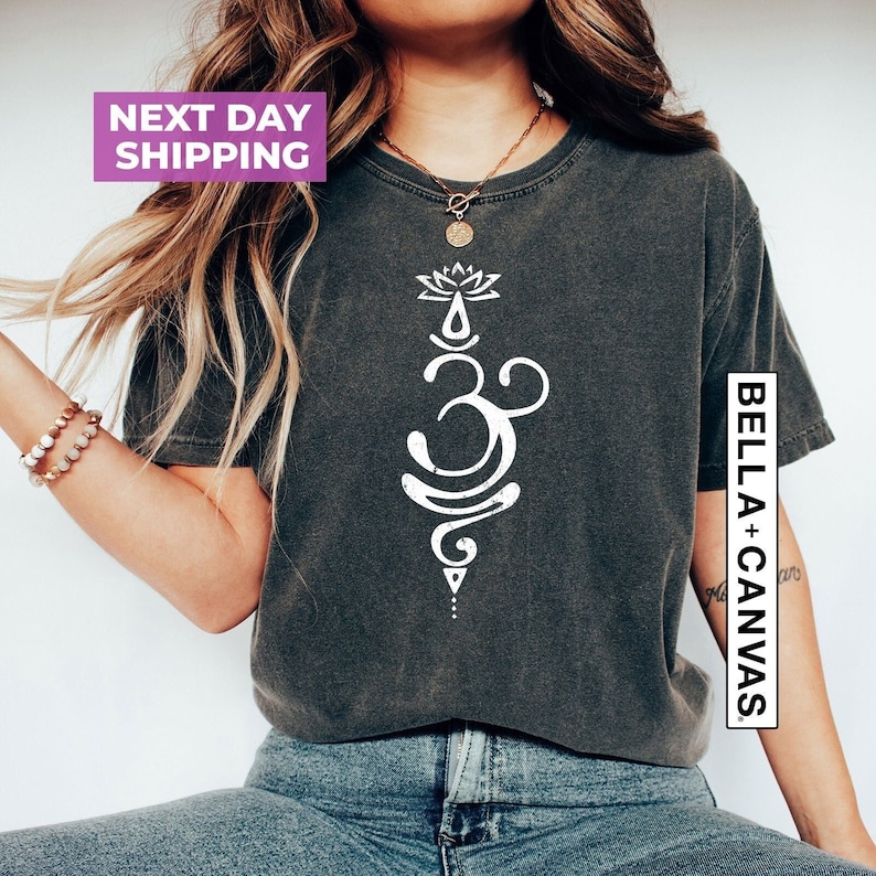 May include: A dark gray t-shirt with a white graphic of an Om symbol with a lotus flower above it. The text "BELLA + CANVAS" is on the right side of the shirt.