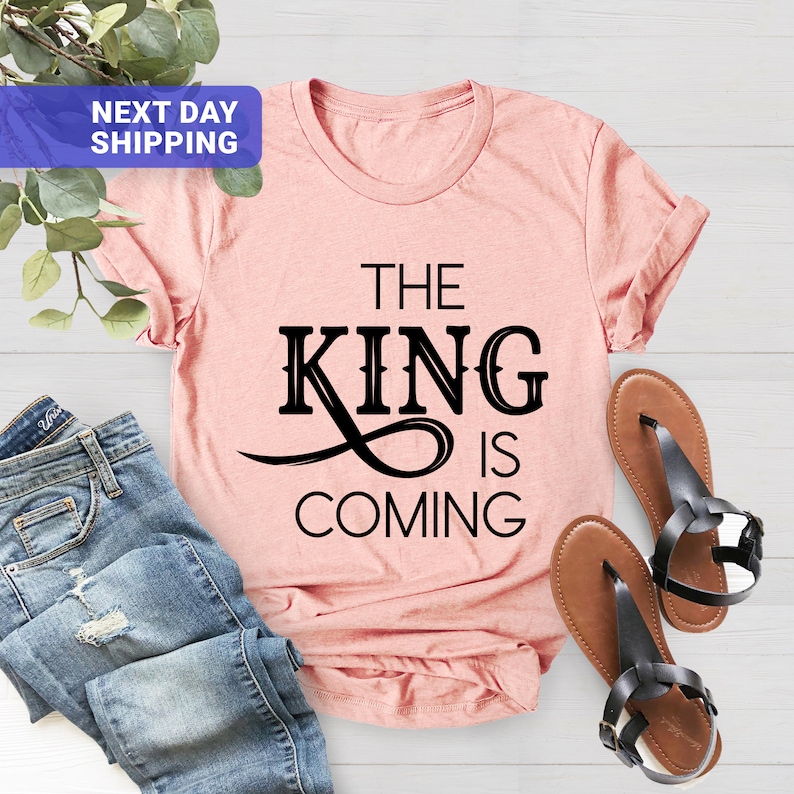 May include: A pink t-shirt with black text that reads "The King is Coming".