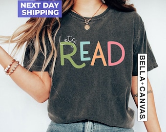 Puede incluir: Una camiseta de manga corta gris carbón con el texto Let's READ en letras multicolores. La palabra Let's está en cursiva blanca, mientras que READ aparece en letras grandes, llamativas y coloridas. Tiene cuello redondo.