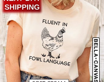 May include: A soft cream-colored short-sleeve t-shirt featuring a black line-art illustration of a chicken in the center. Above the chicken, the text reads FLUENT IN, and below the chicken, the text reads FOWL LANGUAGE.