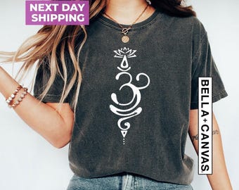 May include: A charcoal gray short-sleeve t-shirt featuring a white vertical graphic of an Om symbol topped with a lotus flower design. The shirt has a crew neckline and a distressed, washed-out fabric texture. The text BELLA+CANVAS is visible.