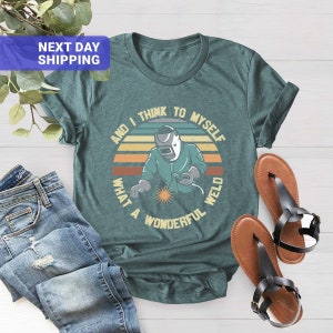May include: A teal blue t-shirt with a retro-style graphic of a welder with the text "And I think to myself what a wonderful weld".