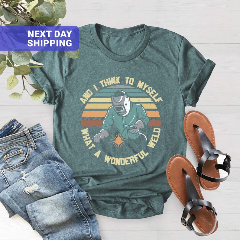 May include: A teal blue t-shirt with a retro-style graphic of a welder with the text "And I think to myself what a wonderful weld".
