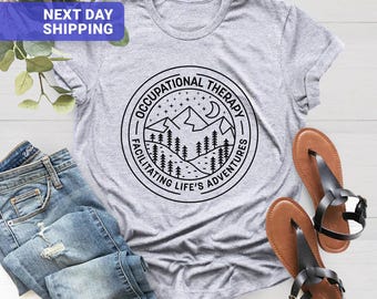 May include: A heather gray t-shirt featuring a black circular graphic with a mountain range, pine trees, stars, and a crescent moon. The text reads OCCUPATIONAL THERAPY FACILITATING LIFE'S ADVENTURES. Blue denim jeans and two brown sandals are also visible.