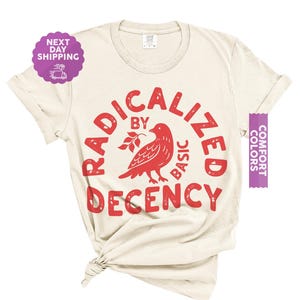 May include: An ivory short-sleeve t-shirt featuring a red graphic print. The design includes a bird illustration with a small branch and the text RADICALIZED BY BASIC DECENCY arranged in a circular pattern around the central bird graphic.