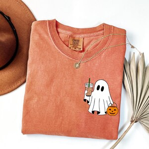 May include: Orange Comfort Colors t-shirt with a round neck. The shirt features a cartoon ghost holding a coffee drink and a jack-o'-lantern. A gold necklace and a brown hat are also in the image. The shirt is a size small.