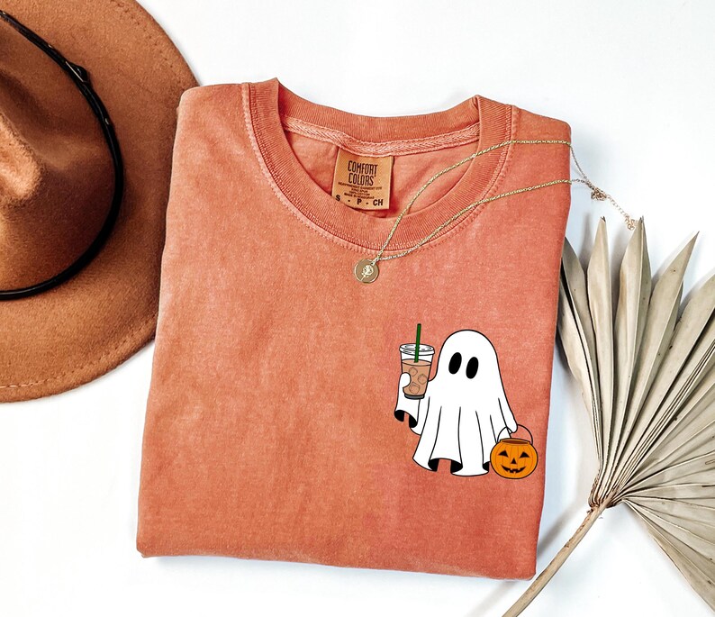 May include: Orange Comfort Colors t-shirt with a round neck. The shirt features a cartoon ghost holding a coffee drink and a jack-o'-lantern. A gold necklace and a brown hat are also in the image. The shirt is a size small.