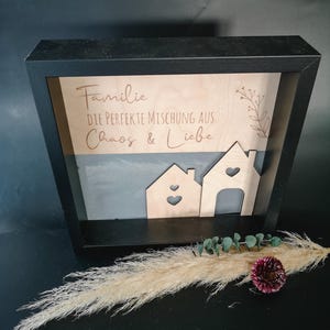 Könnte beinhalten: Ein schwarzer gerahmter Holzschilder mit dem Text "Familie die perfekte Mischung aus Chaos & Liebe" und zwei ausgeschnittenen Holzhäusern mit Herzen. Das Schild ist auf einem grauen Hintergrund.