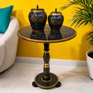 May include: A round pedestal table with a black and gold finish featuring intricate floral patterns. Two matching lidded jars with identical black and gold decorative motifs sit on the tabletop. The table has a central column base.