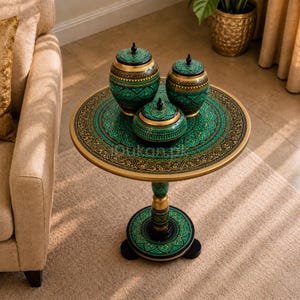 Puede incluir: Una mesa auxiliar redonda con base de pedestal central, con intrincados patrones en turquesa, negro y dorado. Tres tarros con tapa a juego, con motivos decorativos similares en turquesa, negro y dorado, están dispuestos sobre la mesa.