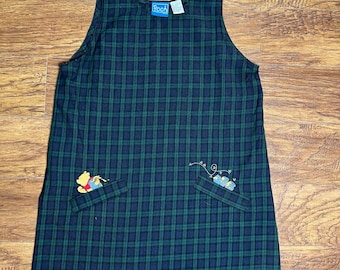 Könnte beinhalten: Grün-marine kariertes Latzkleid mit zwei Taschen. Das Kleid hat Stickereien und ein "Pooh"-Etikett. Knopfverschlüsse an den Trägern. Das Karomuster ist ein klassisches Design.