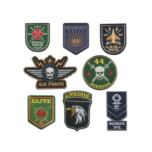 Op de afbeelding: Negen geborduurde patches met militaire thema's. De patches zijn groen, blauw en zwart met gele en rode accenten. De patches bevatten schedels, adelaars, vliegtuigen, kruisen en sterren. De tekst op de patches omvat "Air Force", "Marine", "Special Forces", "Airborne", "Elite", "Division" en "Rescue Div."