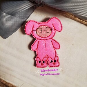 May include: Pink felt applique of a pig wearing glasses and pink pajamas with bunny slippers. The applique is on a light wood background with a gray bow and twine.