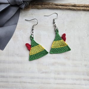 May include: A pair of festive earrings shaped like elf hats. The earrings are green with yellow and red accents, and feature silver-colored ear wires. The earrings are displayed on a white wooden surface with a gray ribbon and twine.
