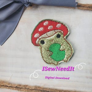 May include: Embroidered iron-on patch featuring a green frog holding a green leaf, wearing a red mushroom hat with white polka dots. The patch has a glittery finish. The text "ISewNeedIt Digital Download" is visible below the patch.