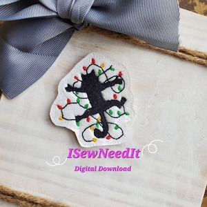 May include: A black cat silhouette tangled in white Christmas lights with red, green, and yellow bulbs. The image is on a white wooden background with a gray bow and twine. The text "iSewNeedIt Digital Download" is below the image.
