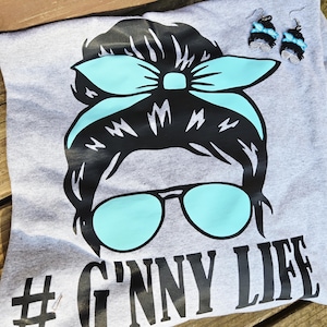 May include: A grey t-shirt featuring a graphic of a woman's face with sunglasses and a headband. The sunglasses and headband are light blue. The text "# G'nny Life" is printed on the shirt. Matching earrings are also visible.